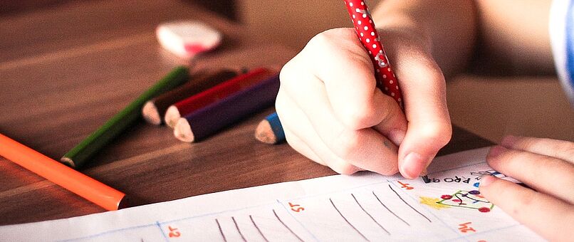 Kind zeichnet und schreibt mit Buntstiften etwas auf ein Papier